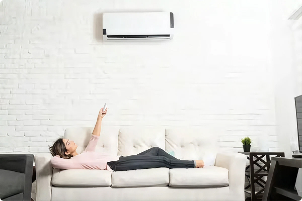 Wall-mounted ductless mini-split head cooling a white brick living room while a woman stretches out on the sofa
