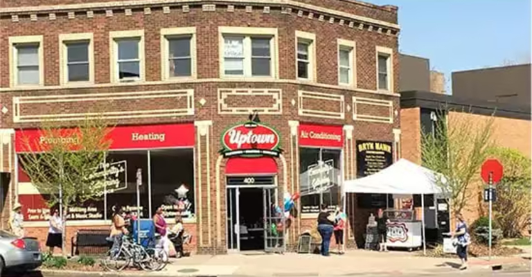Two-story brick corner building with red awnings for plumbing, heating and air conditioning next to the Uptown marquee, with a sidewalk event tent and passersby